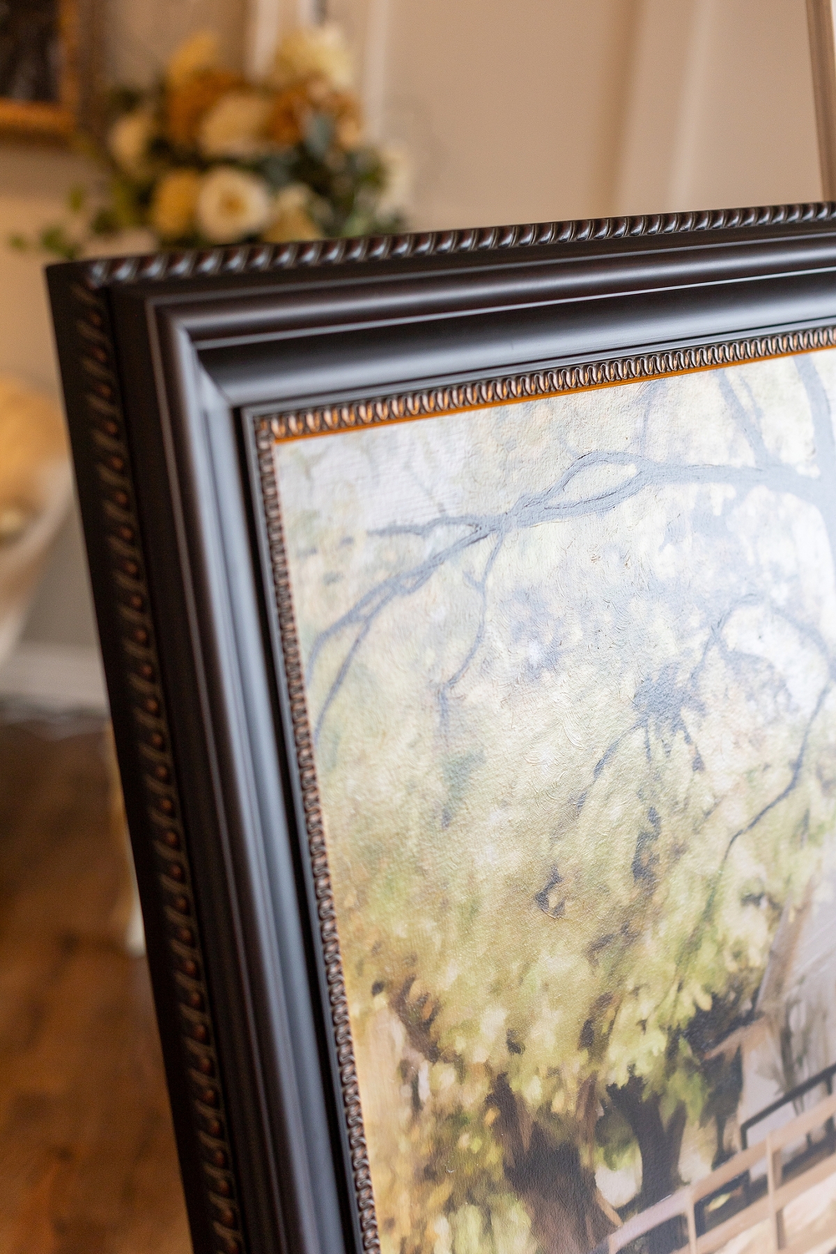 A close-up view of the corner of an ornate picture frame displaying a painting with visible tree branches and foliage.
