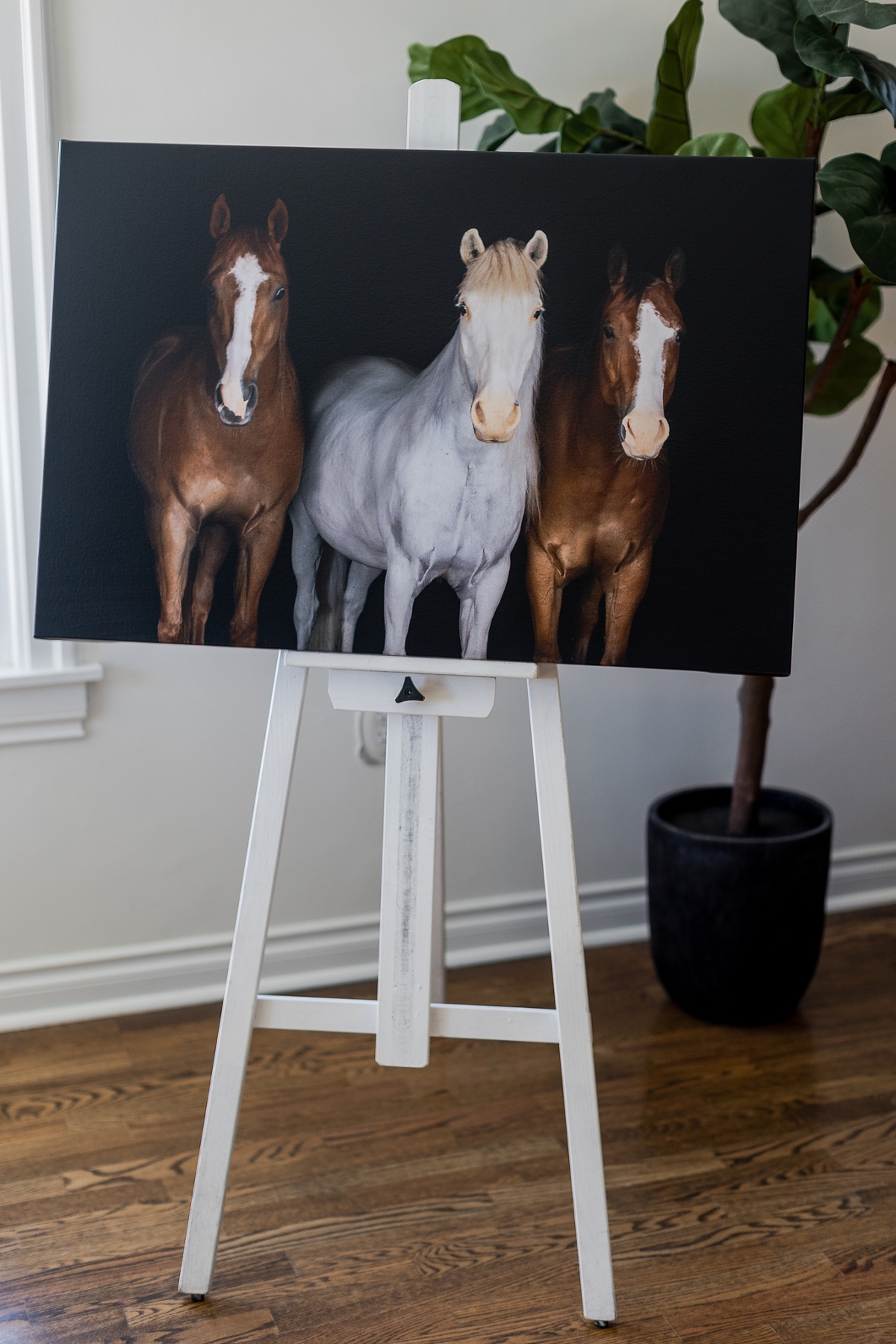 A painting of three horses displayed on a white easel, with a potted plant in the background.