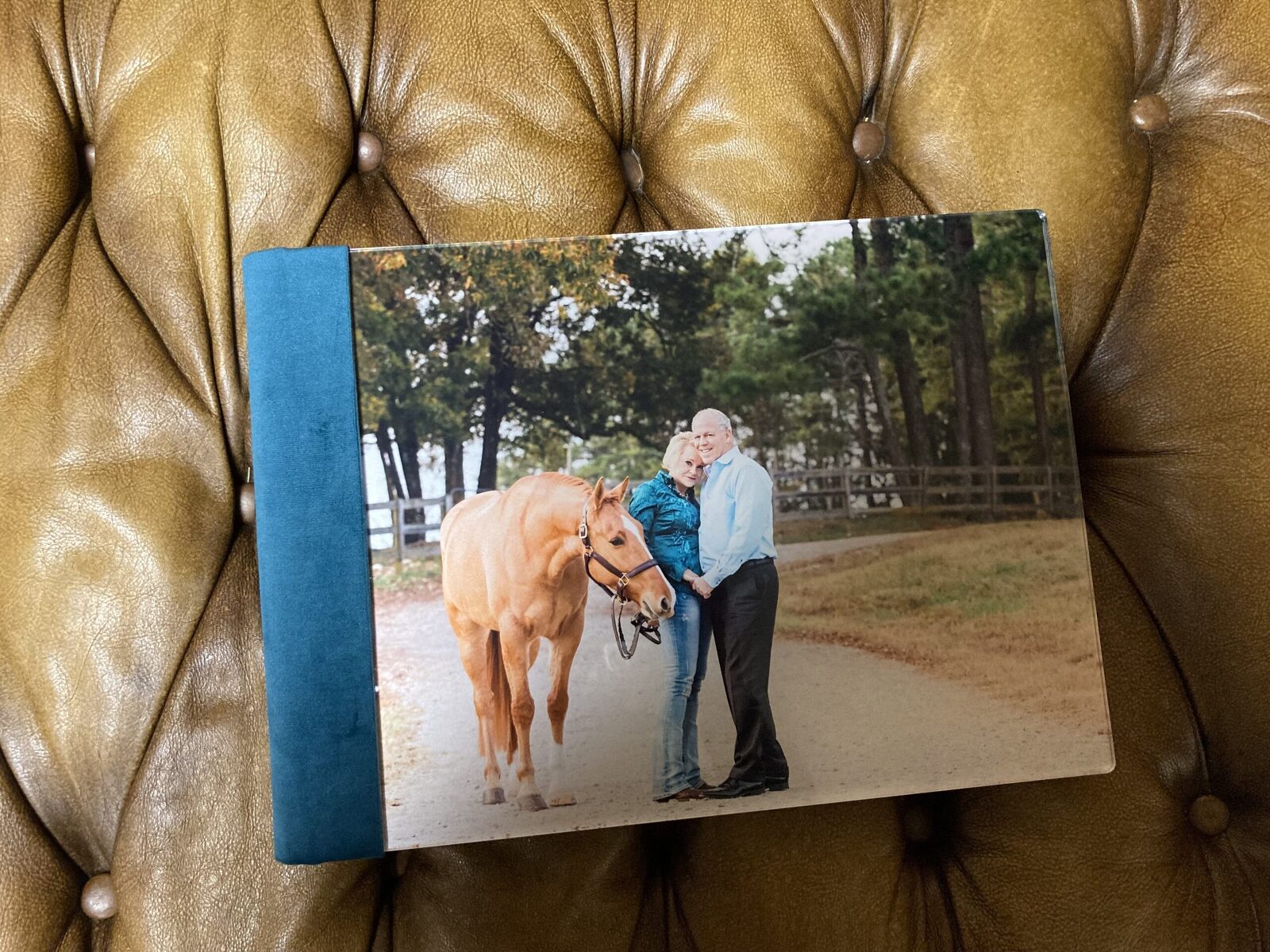 A photo album on a leather surface shows a couple standing with a brown horse on a dirt path lined with trees.